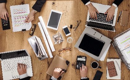 Overhead image of people working on a long desk with laptops, calculators, pens and books.